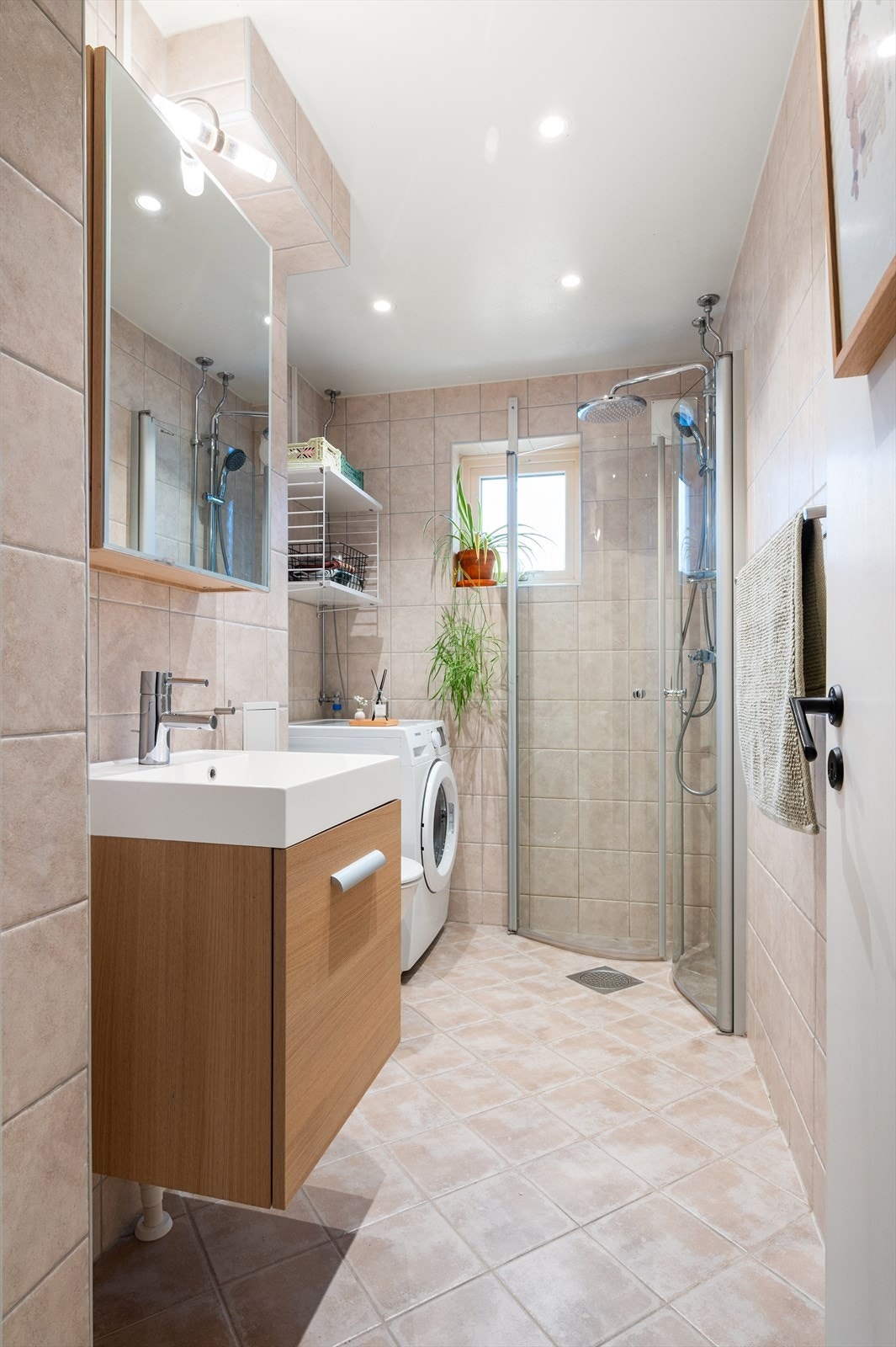 Badet inneholder gulvstående toalett, servant på vegghengt servantskap med speil og lys over. Dusjhjørne med vegghengte dursjører. Galleribilde