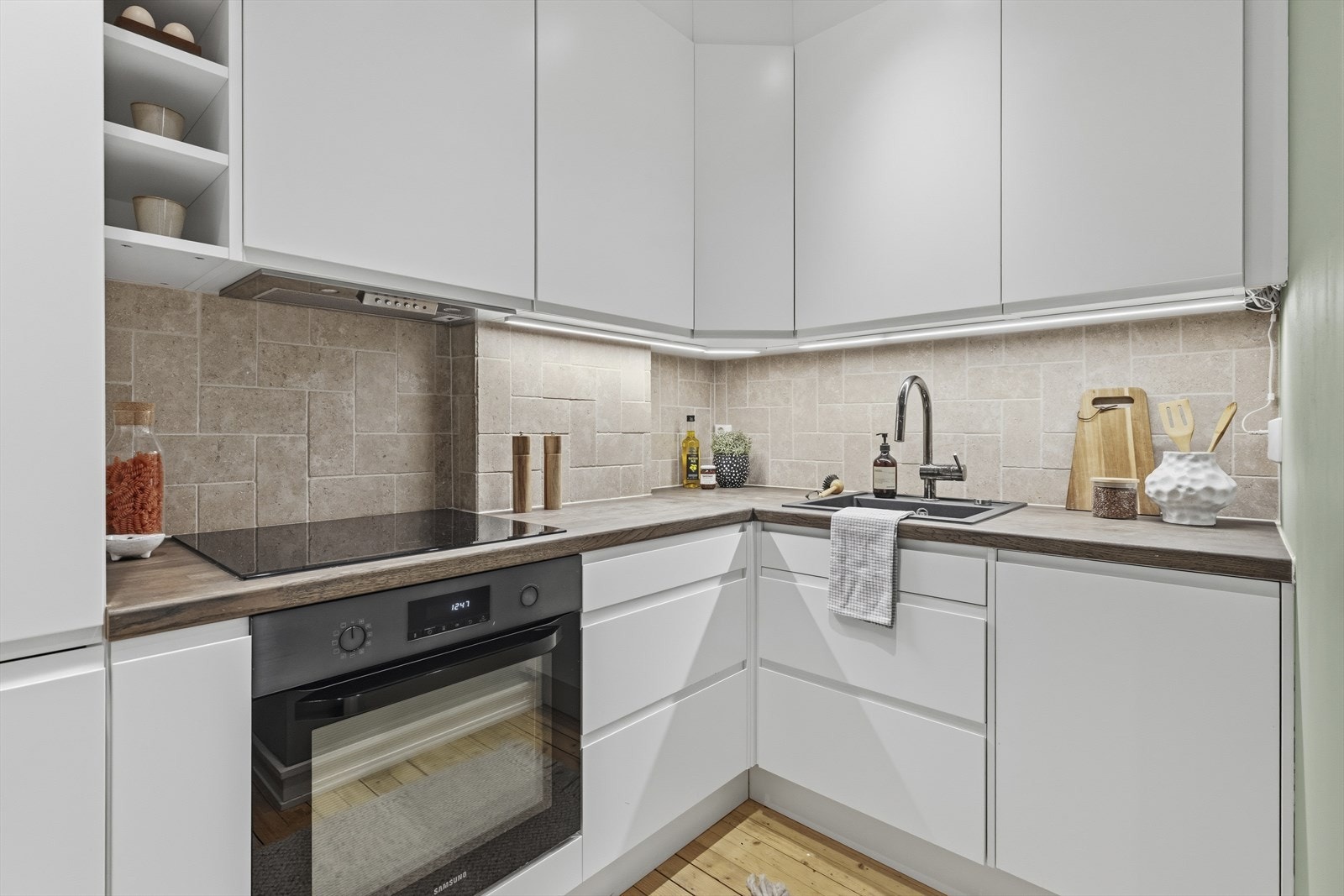 Benkeplaten i tre skaper en varm kontrast til den lyse kjøkkeninnredningen, og flislagt vegg over benk gir et praktisk og dekorativt arbeidsområde. Kjøkkenet er utstyrt med integrerte hvitevarer, herunder kjøleskap med frysedel, induksjonst Galleribilde