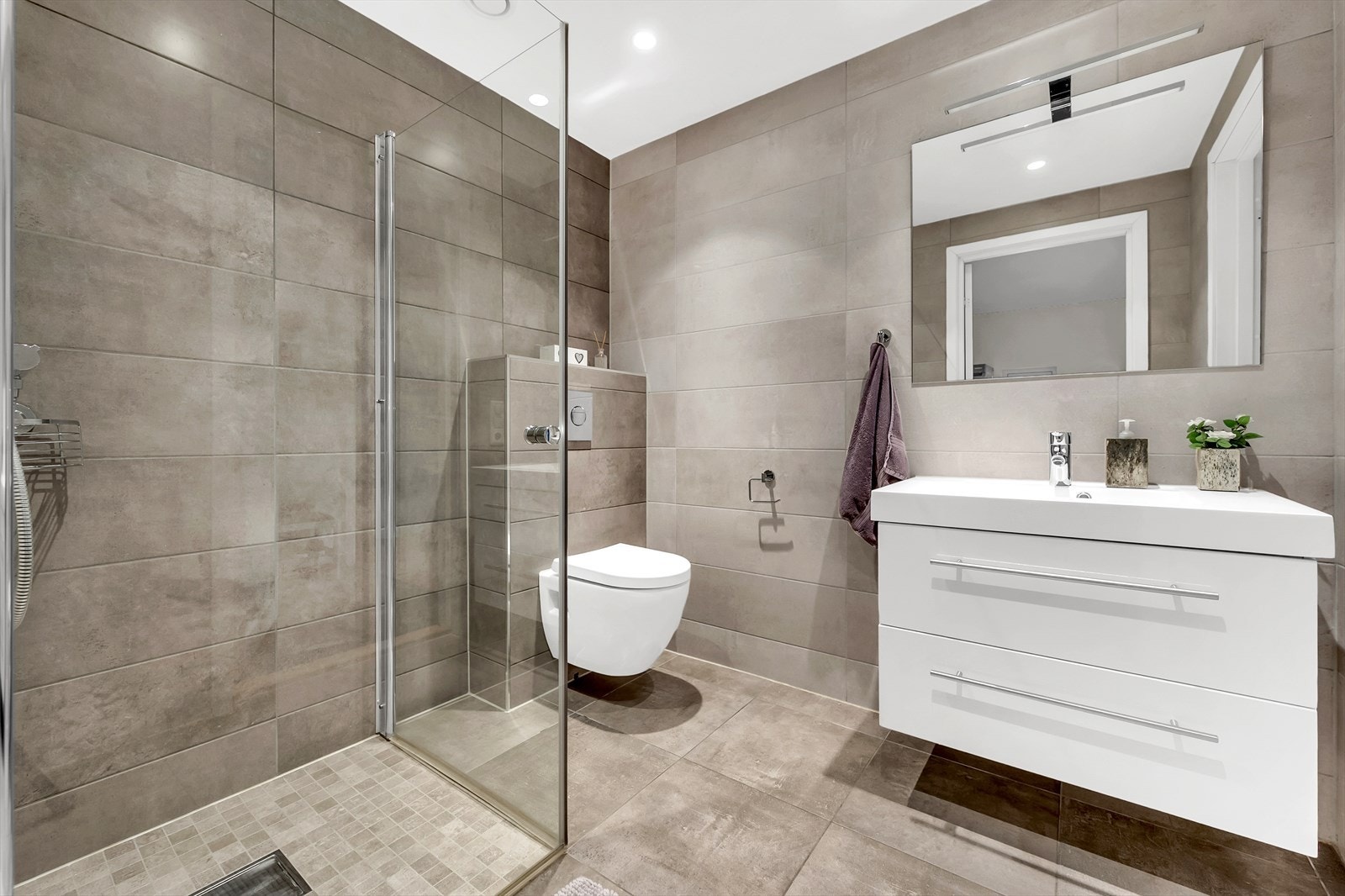 Delikat baderom med dusjhjørne, toalett og servant med oppbevaring. Beige fliser gir en god romfølelse. Galleribilde