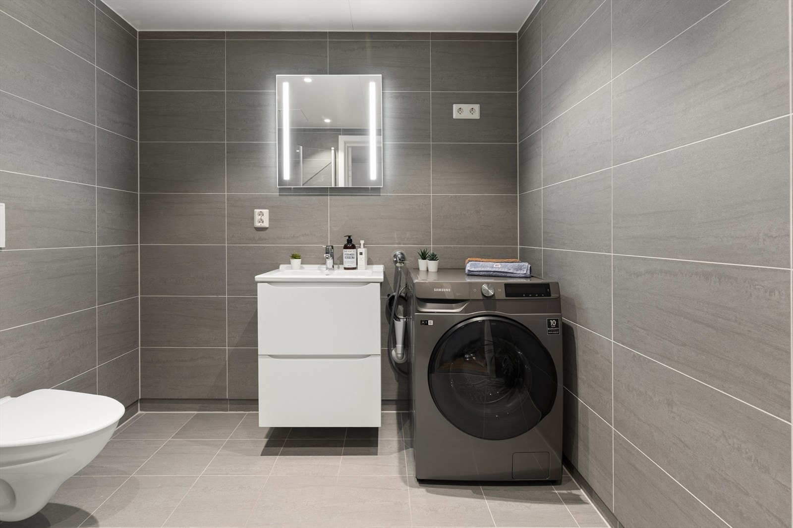 Flislagt bad med vegghengt toalett, servantinnredning og opplegg for vaskemaskin. Galleribilde