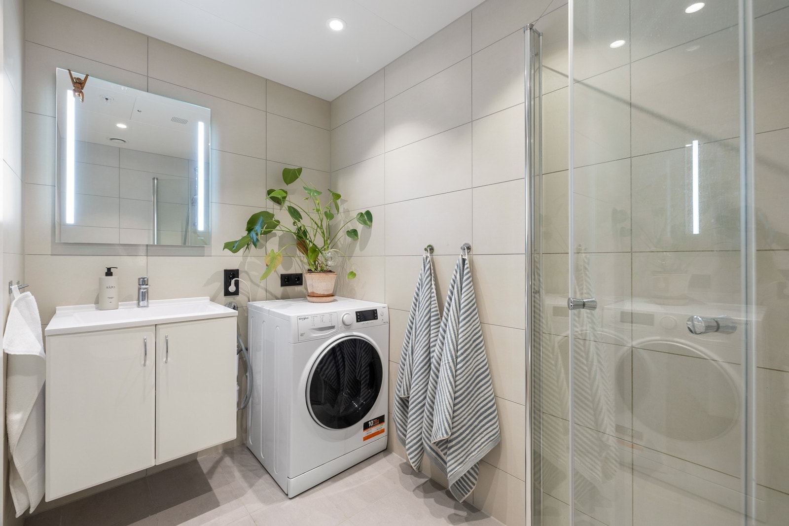 Badet inneholder innfellbare og plassbesparende dusjdører i glass, dusjbatteri og garnityr montert på vegg, kran og avløp til vaskemaskin og veggmontert WC. Rørskapet er lokalisert i himling. Galleribilde