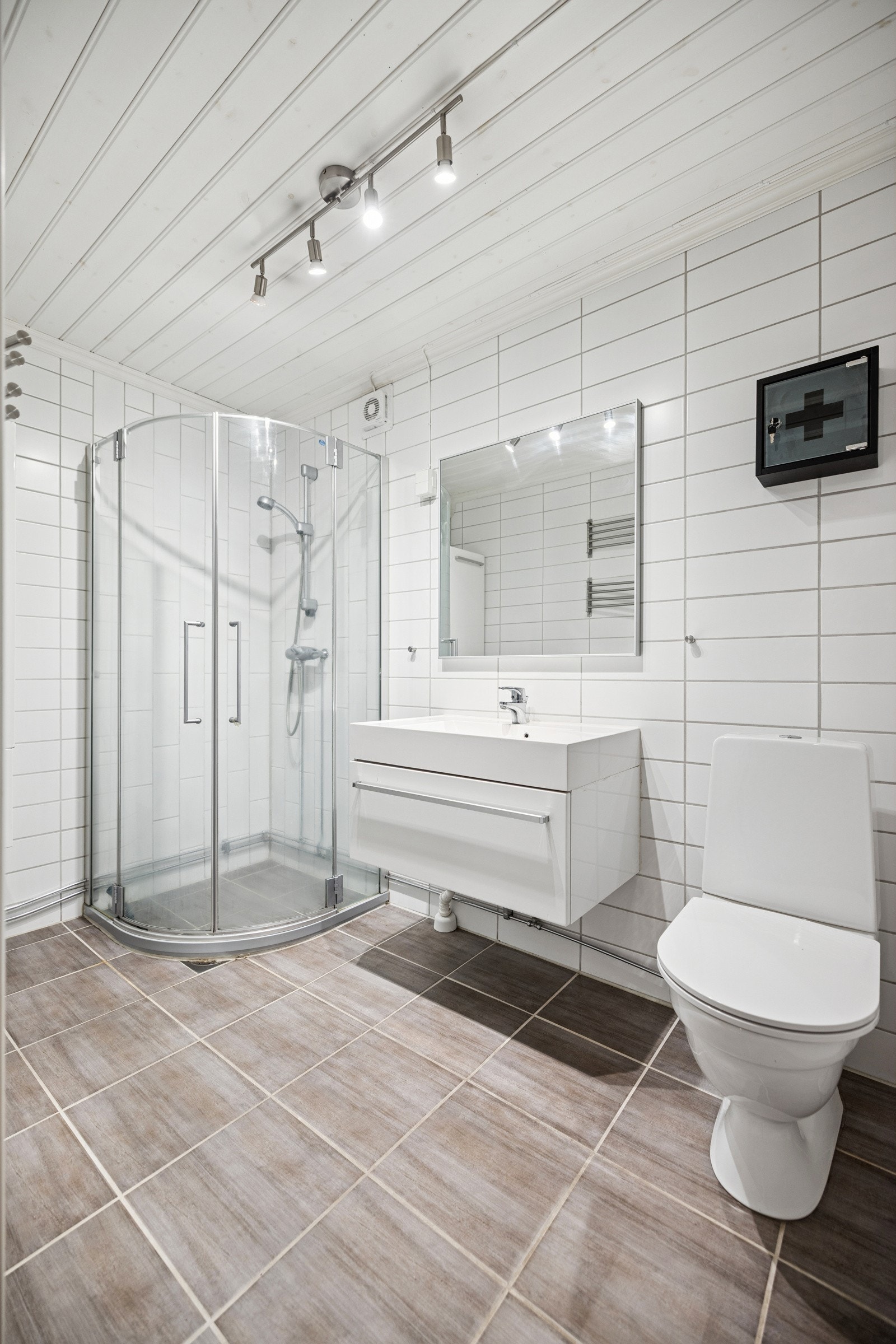 Badet har varmekabler under flislagt gulv og plater i flisemønster på vegg. Innredning med heldekkende servant på underskap og speil over. I tillegg er det montert langskap på vegg, dusjnisje og wc. Galleribilde