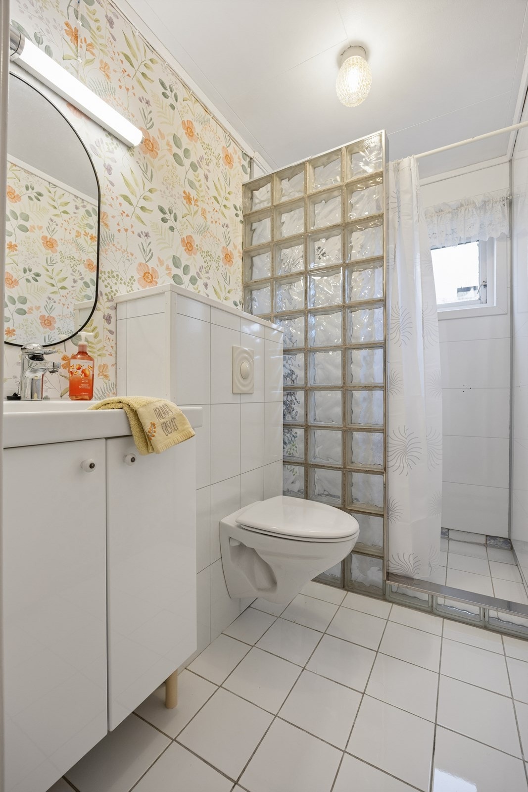 Badet er innredet med hvite fliser på gulvet og en kombinasjon av baderomsplater og tapetserte veggflater. I gulvet har man også varme for ekstra komfort. Badet er ellers utstyrt med dusjhjørne, vegghengt toalett og baderomsinnredning med nedfelt servant. Galleribilde