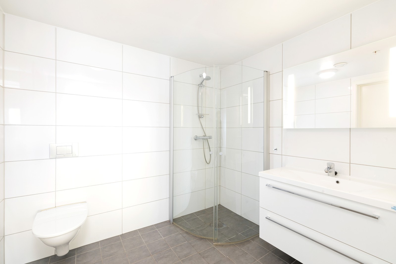 Flott flislagt bad med varme i gulv. Badet har dusjhjørne, vegghengt toalett og servant med underskuffer og overspeil med lys. Galleribilde