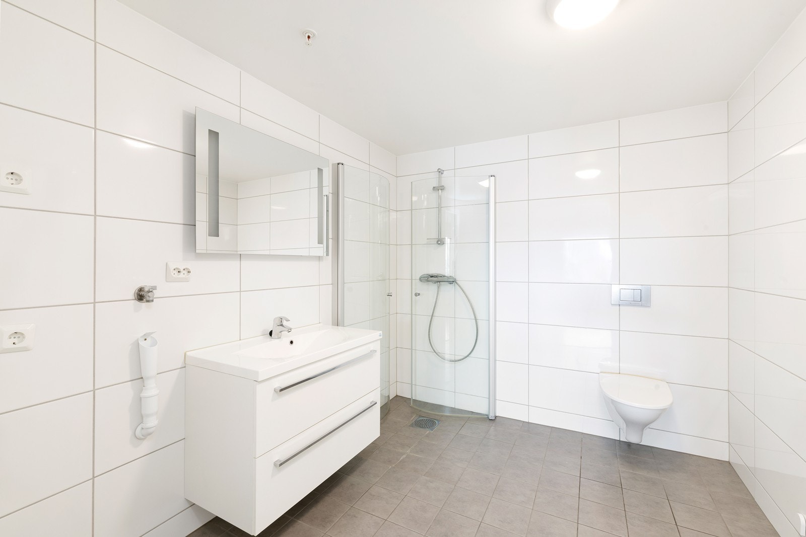 Badet har dusjhjørne, vegghengt toalett og servant med underskuffer og overspeil med lys. Galleribilde