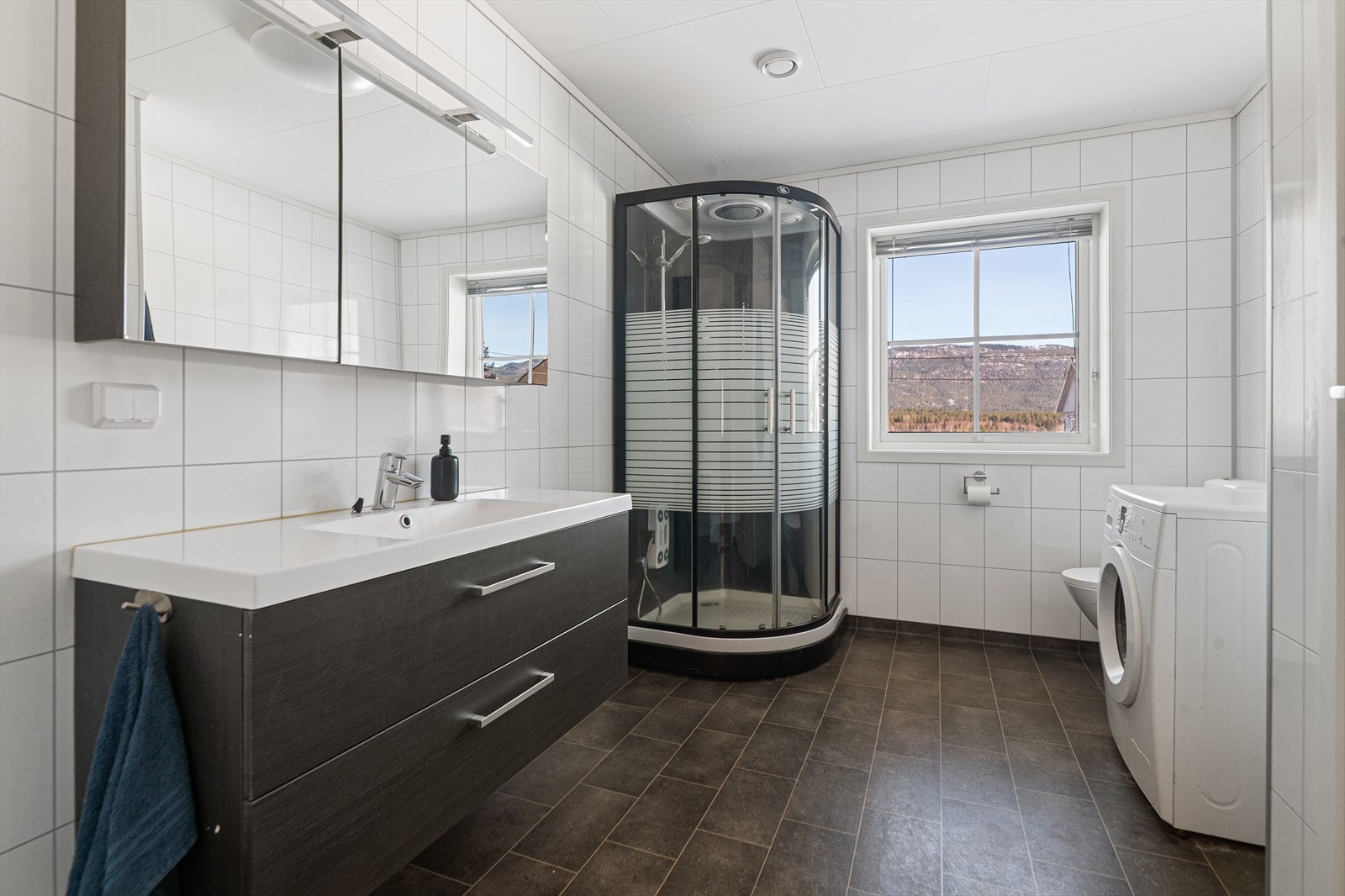 Lyst og pent bad fra byggeår som inneholder heldekkende servant med underskap, speilskap med lys, gulvmontert toalett, dusjkabinett og opplegg til vaskemaskin. Galleribilde