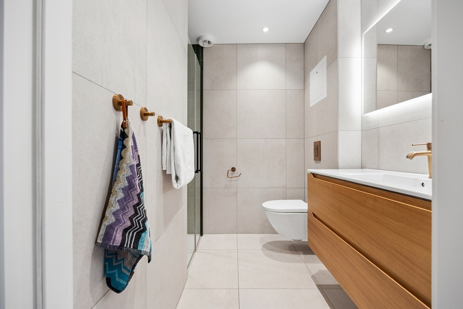 Lekre, store fliser, varmekabler i gulv, downlights i tak, vegghengt toalett, speil med belysning, samt en pen baderomsservant Galleribilde