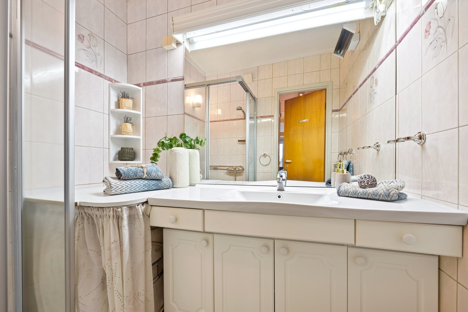 Badet har innredning med hvit innredning med profilerte fronter og servant. Speil med lys og skaper under servant. Fliser på vegger og gulv. Galleribilde