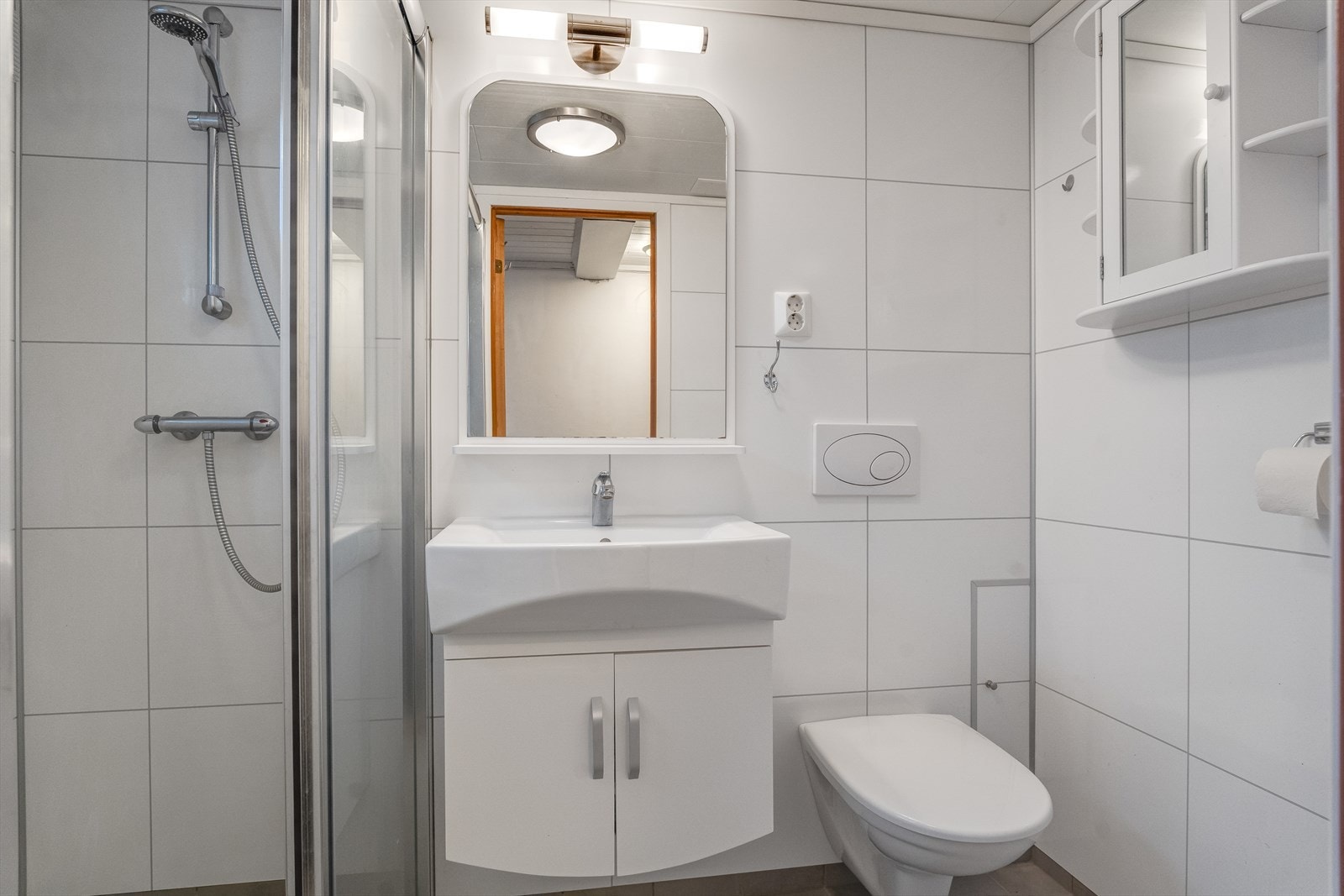 Bad med fliser på gulv og baderomsplater på vegger, vegghengt toalett, dusj og baderomsinnredning med skuffer og speil. Galleribilde