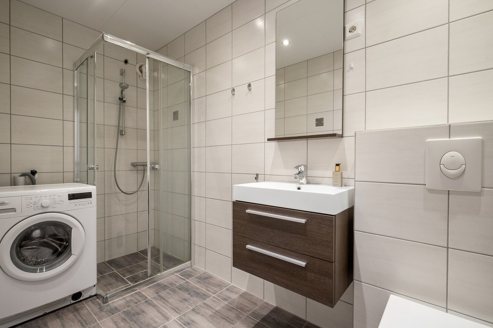 Badet har fliselagt gulv og elektriske varmekabler som oppvarming, og innredet med toalett, servant, vegghengt toalett, vaskemaskin og dusjhjørne Galleribilde