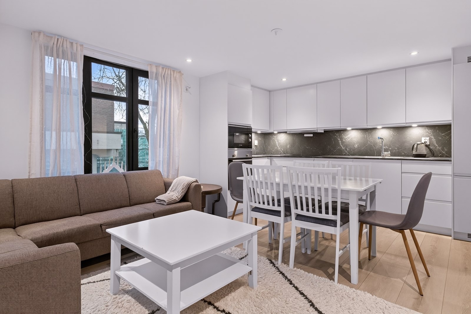 Lys og luftig stue i åpen løsning mot kjøkken, med store vinduer og spotter som gir et flott og jevnt lys gjennom hele rommet. Galleribilde