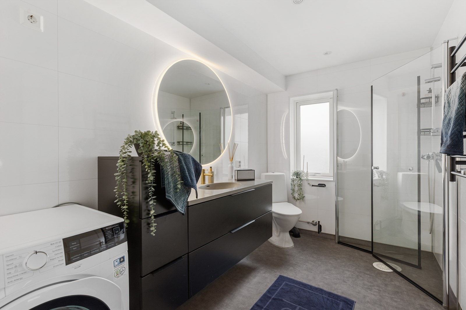 Lyst og innbydende bad med tidløse overflater og elektrisk gulvvarme. Badet har servantinnredning, gulvmontert toalett, dusjhjørne og opplegg for vaskemaskin. Galleribilde