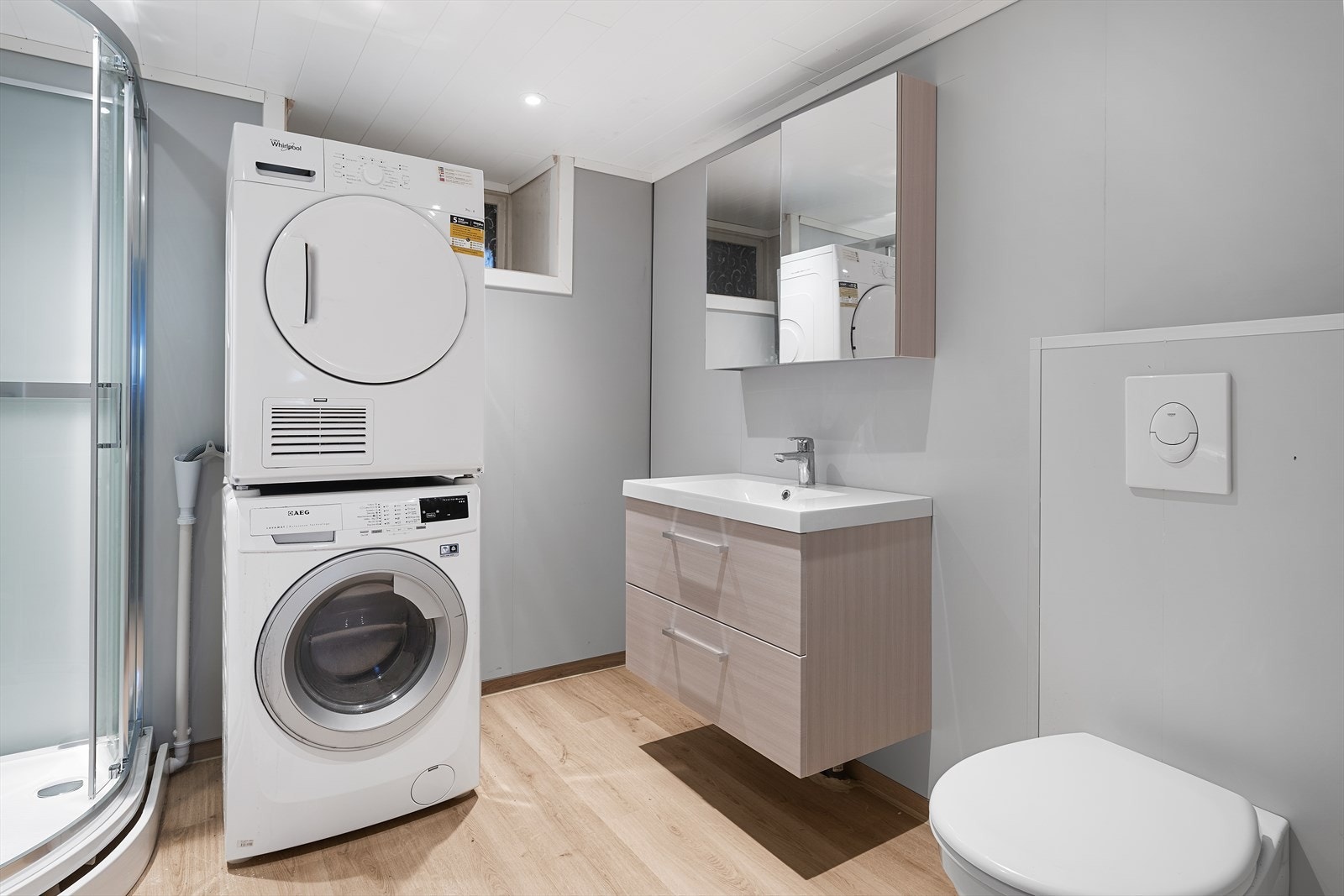 Lyst baderom med klikk-vinyl på gulv. Badet er utstyrt med servant over skuffeinnredning, speilskap, dusjkabinett og vegghengt toalett. Galleribilde