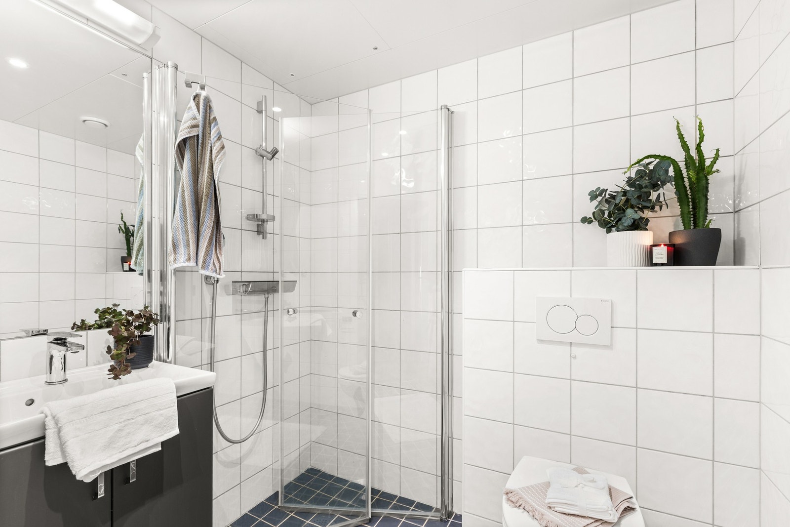 Badet har et dusjhjørne med glassdører og dusj rett på flis, veggmontert servant, elektrisk håndkletørker, toalett og opplegg for vaskemaskin Galleribilde