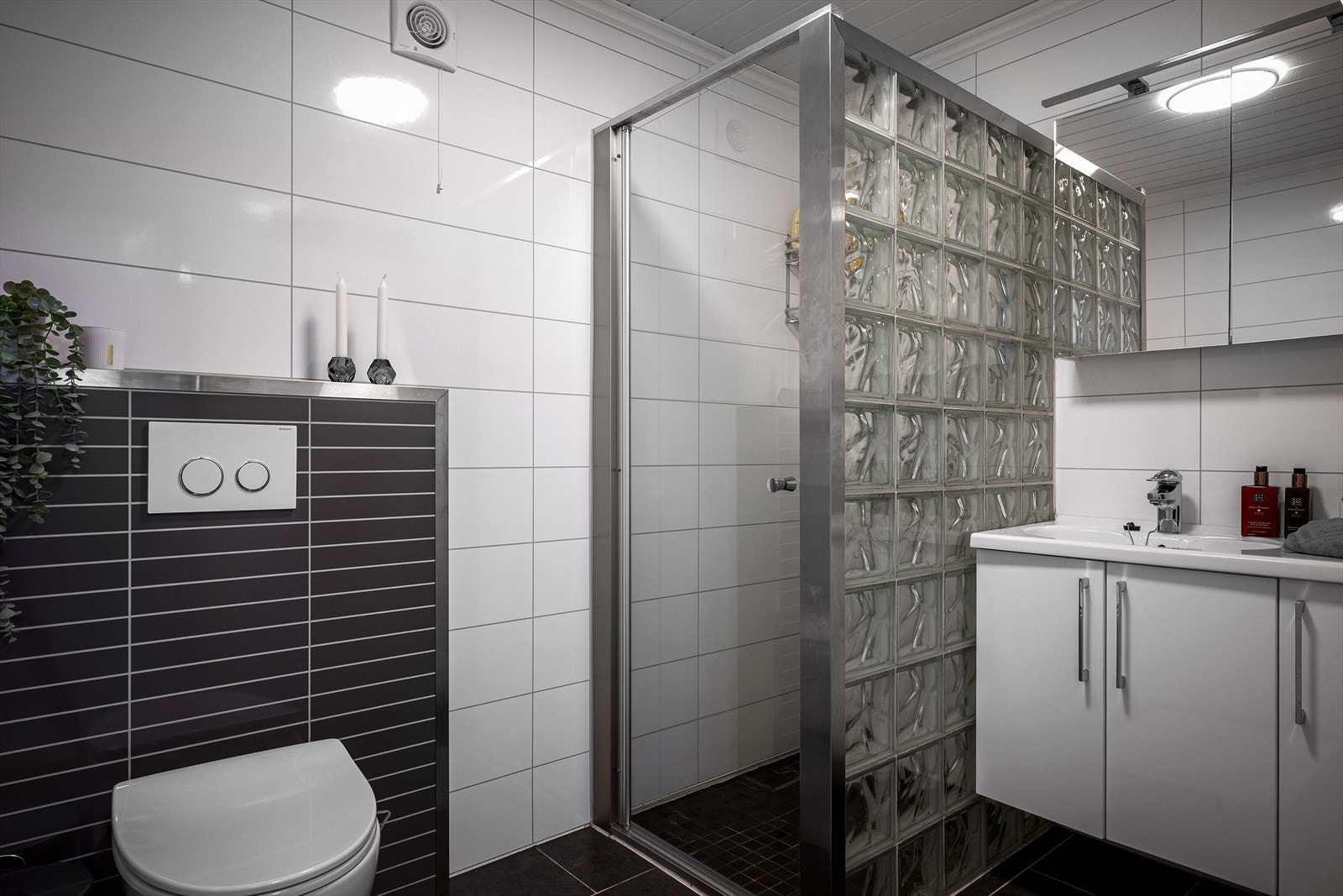 Boligens bad er elegant med et flislagt gulv i mørk tone og hvite baderomsplater på veggene. Rommet har et stilrent uttrykk med innredning som inkluderer nedfelt servant, et veggmontert toalett og dusjdører. Dusjens glassbyggerstein tilfører et eget preg. Galleribilde