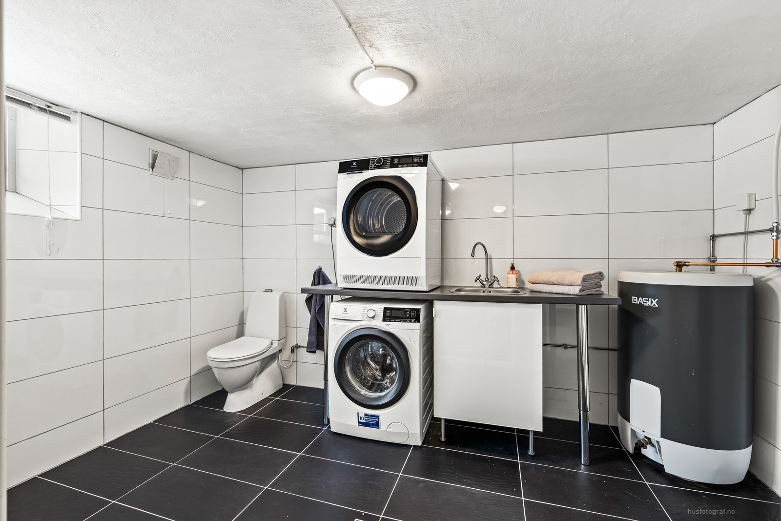 Stort, flislagt vaskerom med toalett, opplegg for oppvaskmaskin og praktisk utslagsvask med benk Galleribilde