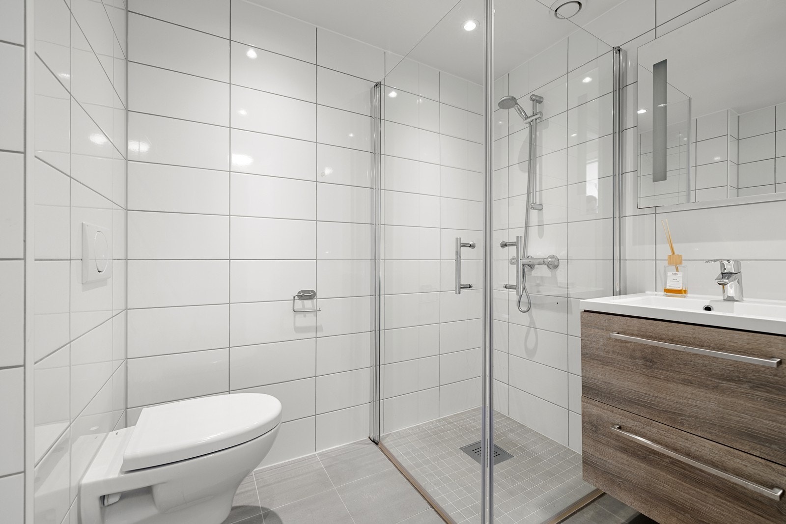 Delikat baderom med flislagte overflater. Badet er utstyrt med vegghengt toalett, servant med skuffer og dusjnisje med innfellbare glassdører. Galleribilde