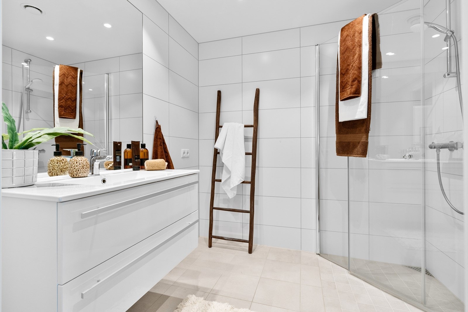 Badet er flislagt med beige fliser på gulv og hvite fliser på veggene. Her finner du et stort servantskap med god oppbevaringsplass, speil, toalett og et praktisk dusjhjørne. Galleribilde