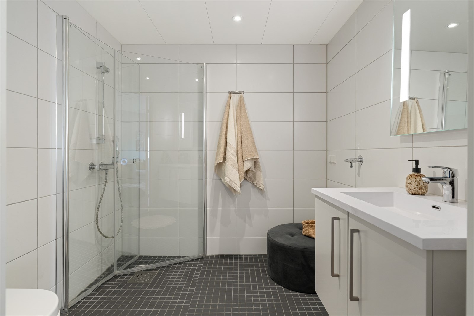 Badet har flislagte overflater, gulvvarme og downlights i himling. Øvrig innredning består av dusjnisje, toalett, opplegg for vaskemaskin og en lys baderomsinnredning med god lagringsplass. Galleribilde