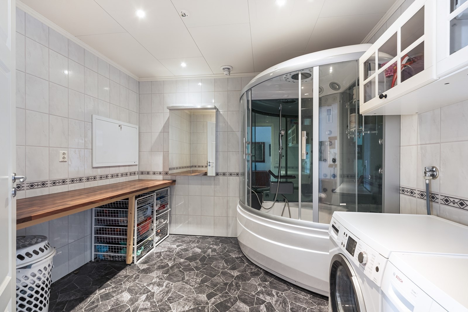 Praktisk vaskerom hvor det er montert benkeplate og overskap. Rommet er utstyrt med vannbåren gulvvarme og et dusjkabinett med badekar. Galleribilde