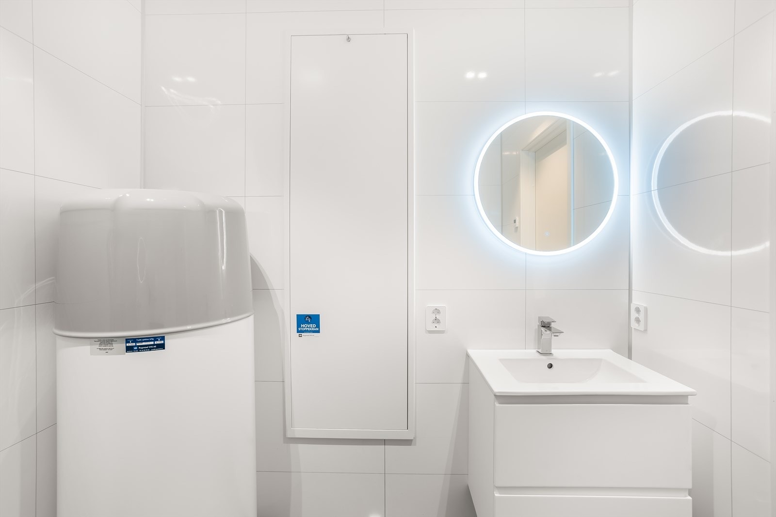 Baderomsinnredning i hvit høyglans, vegghengt toalett og speil med integrert belysning. Det er også opplegg for vaskemaskin. Galleribilde