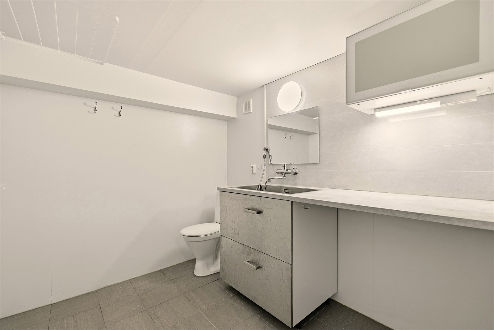 Toalettrommet inneholder innredning med slette fronter, benkeplate i laminat og nedfelt stålvask med vegghengt blandebatteri, speil og gulvstående toalett. Rommet har varme i gulv og klaffeventil på vegg. Galleribilde