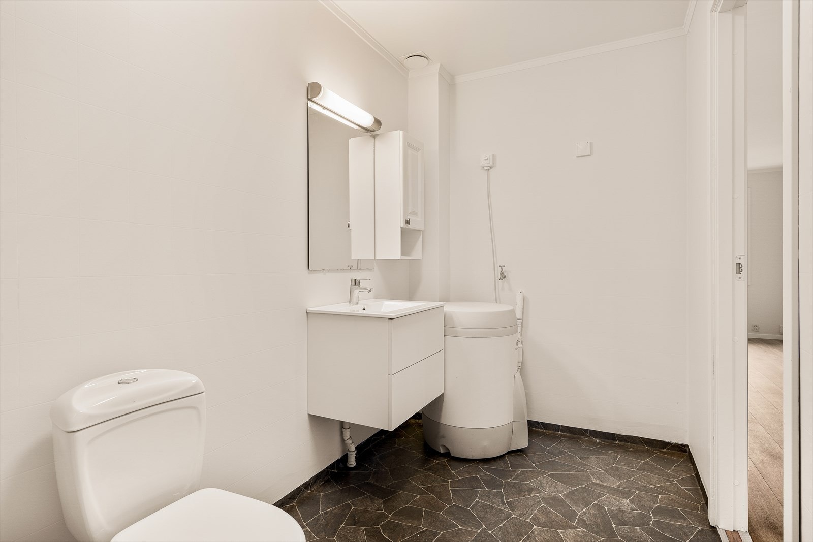 Badet er kombinert vaskerom med vinylbelegg på gulv, servantinnredning og opplegg for vaskemaskin. Galleribilde