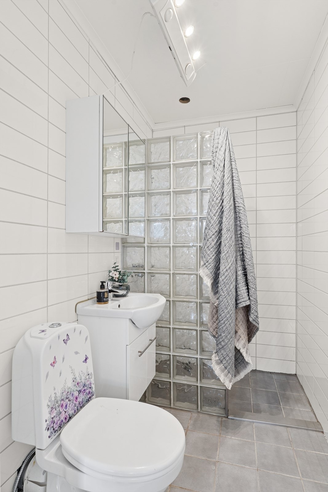 Bad 2: Eldre dusjbad utstyrt med en dusjnisje med vegg av glassbyggerstein, en servant med skapinnredning og et toalett. Galleribilde