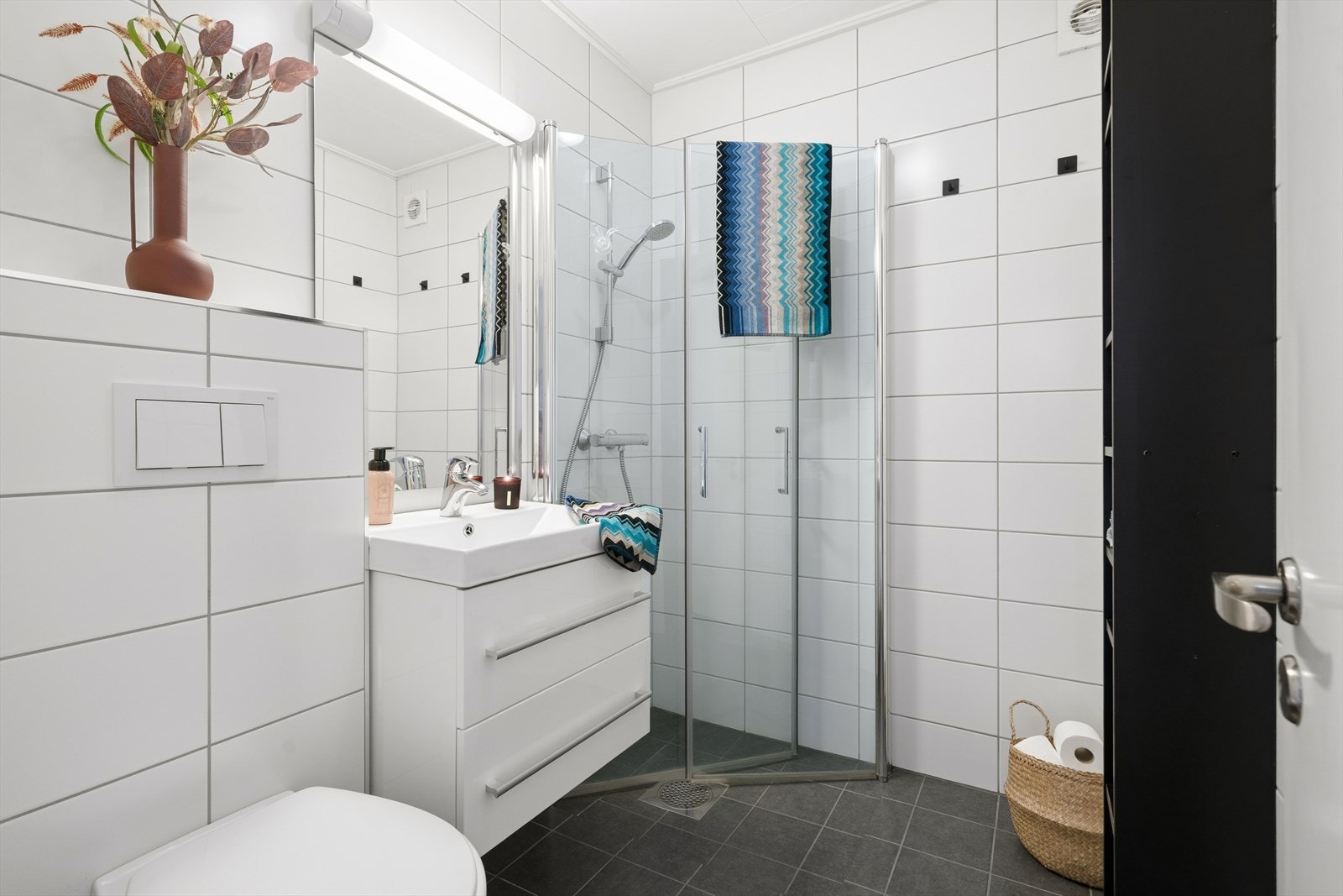 Lekkert og velholdt bad med moderne overflater og gjennomførte materialvalg. Rommet har store, hvite fliser på veggene som gir et tidløst og rent uttrykk, kombinert med mørke gulvfliser som tilfører en elegant kontrast Galleribilde