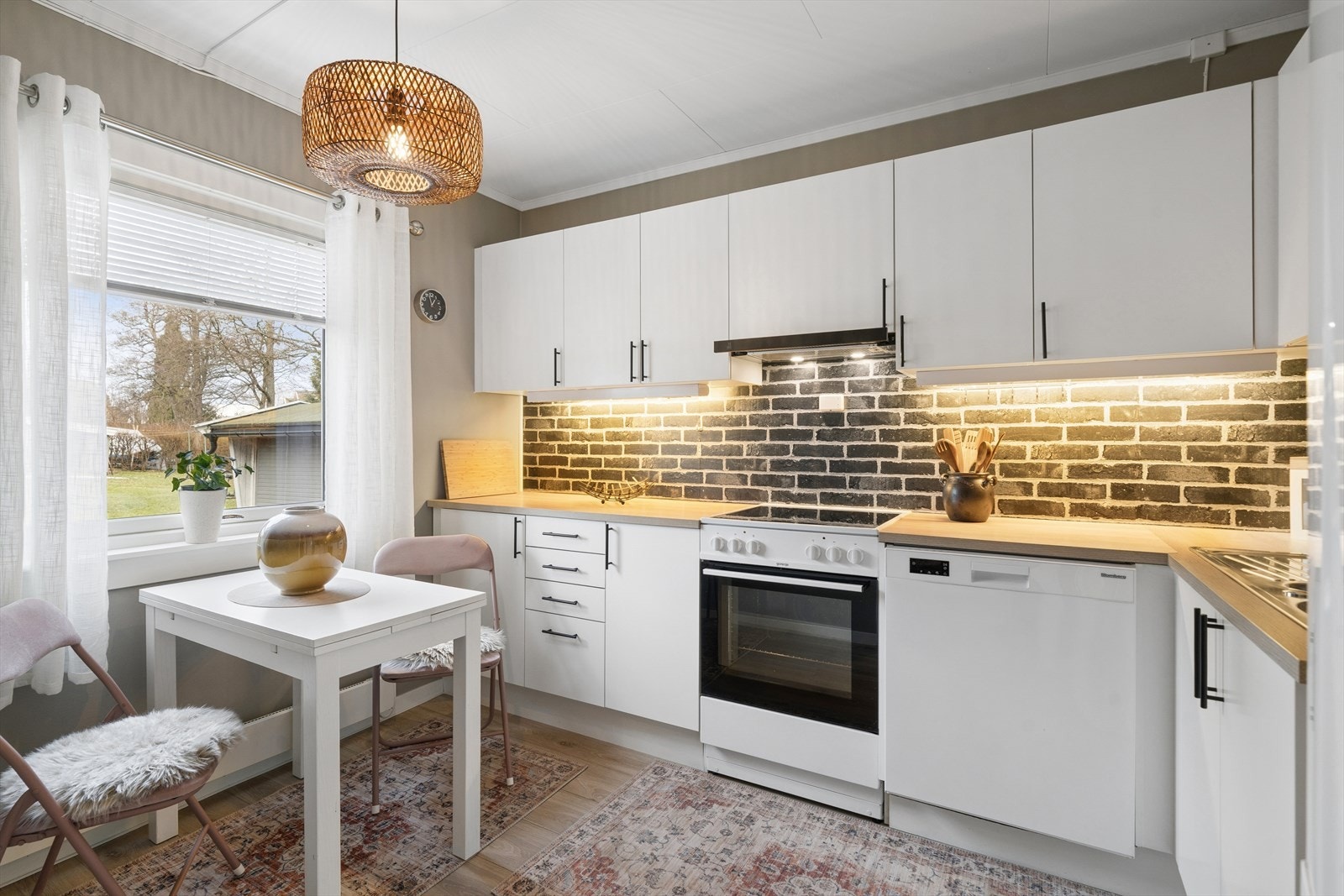 Moderne og innbydende kjøkken med pene, hvite fronter og laminerte benkeplater som gir en varm og naturlig kontrast. Integrert belysning under overskapene sørger for godt arbeidslys og en behagelig atmosfære. Galleribilde