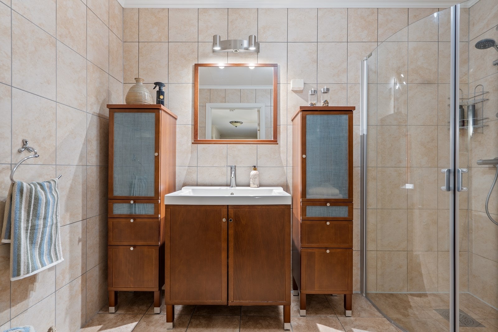 Romslig helfliset baderom innredet med wc, vask i seksjon med tilhørende oppbevaring i skap samt dusjhjørne. Galleribilde