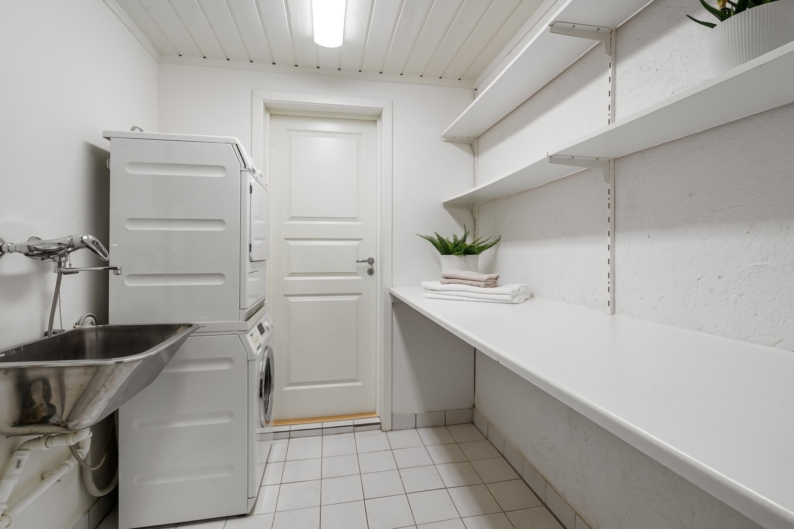 Praktisk vaskerom med hvite overflater, gulvvarme, vegghengte hyller, utslagskum og opplegg for vaskesøyle. Galleribilde