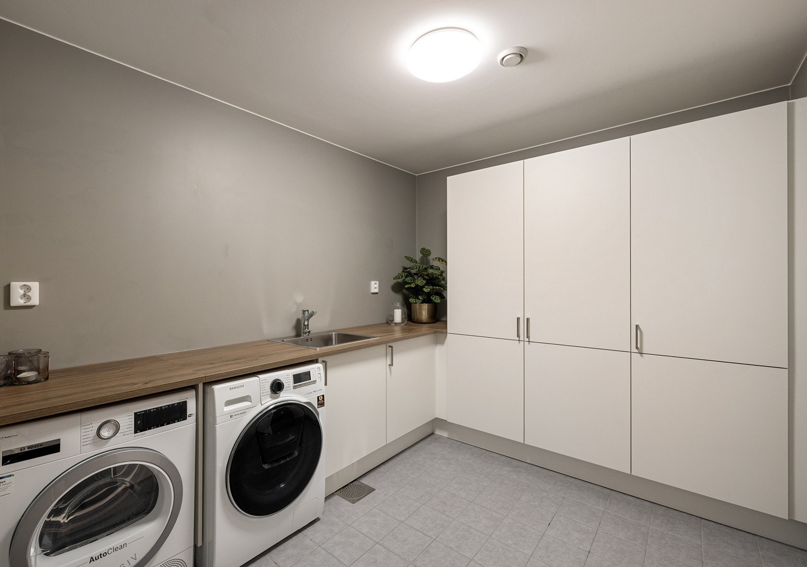 Praktisk og romslig vaskerom med flislagt gulv med gulvvarme og glatte veggflater. Rommet er innredet med underskap, høyskap og utslagsvask, som gir gode oppbevaringsmuligheter og arbeidsflater. Galleribilde