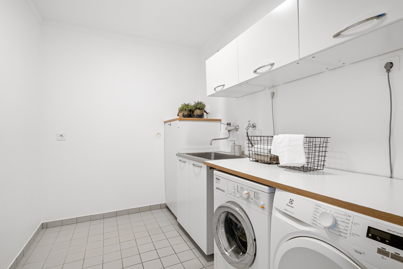 Meget praktisk og romslig vaskerom med rikelig plass til oppbevaring i skap på vegg.Opplegg for vaskemaskin og tørketrommel, som er plassert ved siden av hverandre og med benkeplate over. Utslagsvask. Det er fliser på gulv og slette vegger. Galleribilde