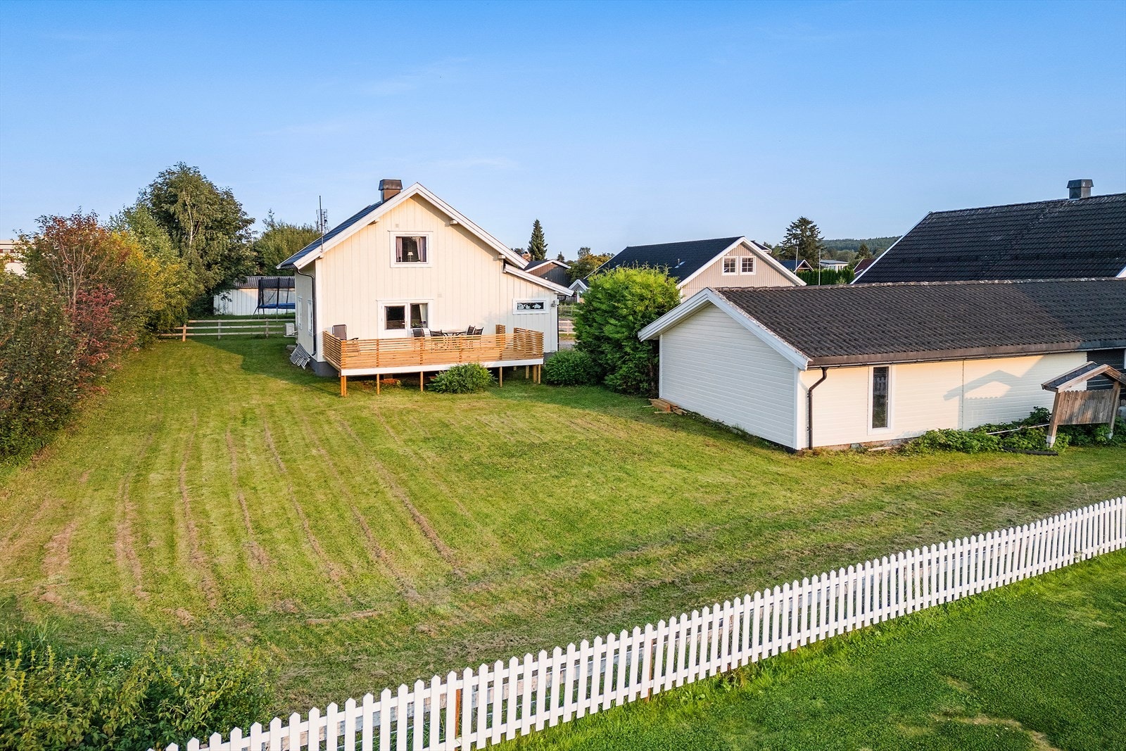 Stor og velstelt hage med grønn plen, hvitt stakittgjerde og en koselig treterrasse tilknyttet huset. Et perfekt sted for lek, grilling og avslappende sommerdager under åpen himmel. Galleribilde