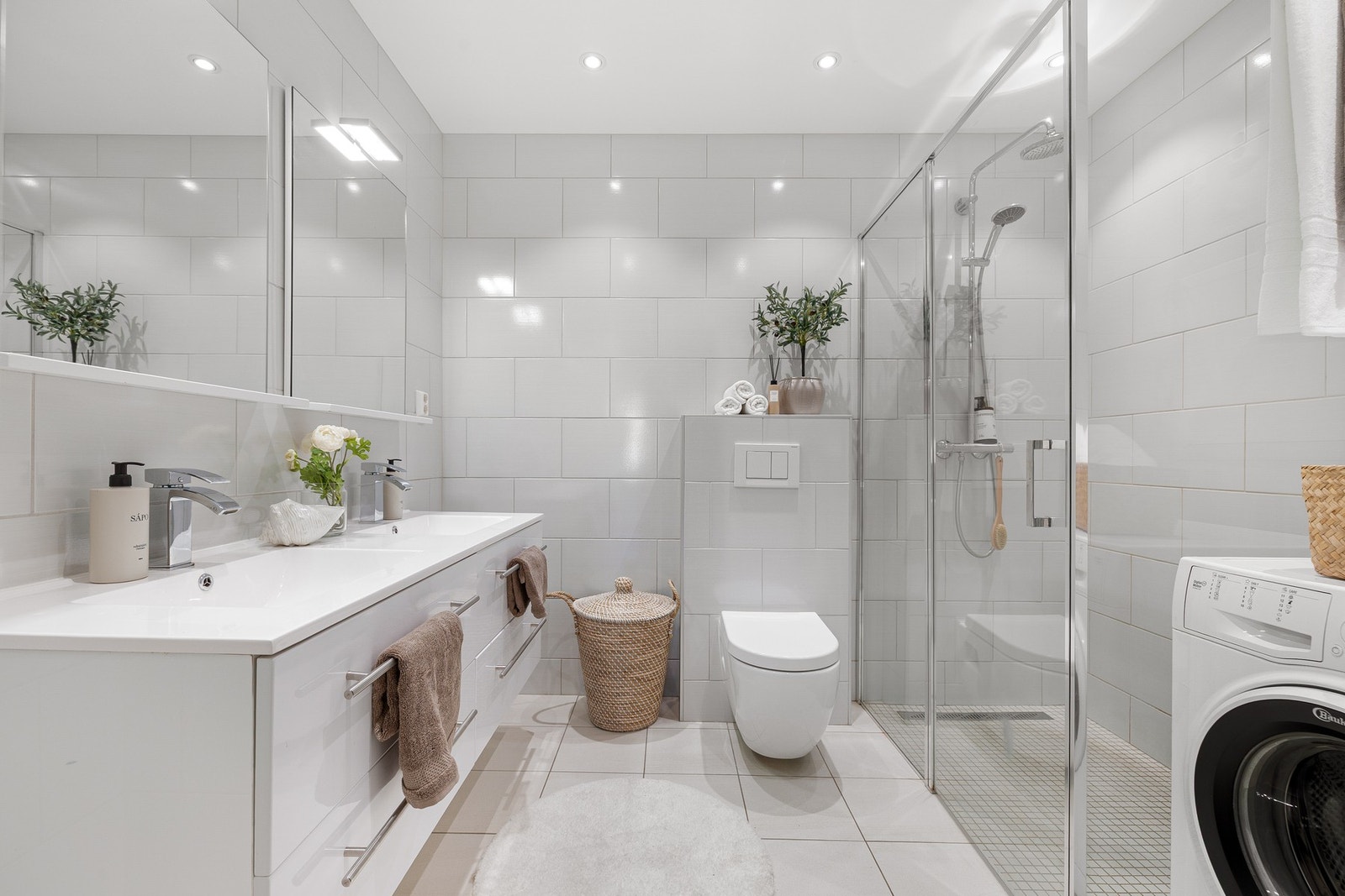 Det lekre baderommet fra 2018 med fliser på både gulv og vegger fremstår innbydende og lyst. Her er det kran og avløp til vaskemaskin, dusjvegg og dusjdør i glass, dobbel servant, speil og veggmontert WC. Galleribilde