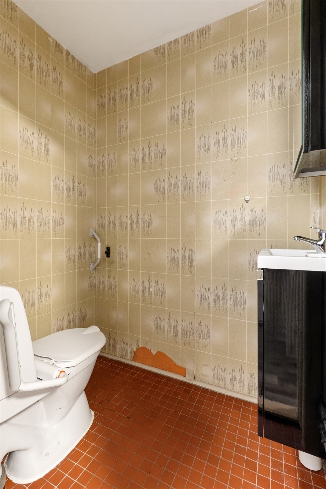 Eldre toalettrom med flislagt gulv, tapet på veggene og enkel innredning bestående av toalett og servant med underskap. Galleribilde