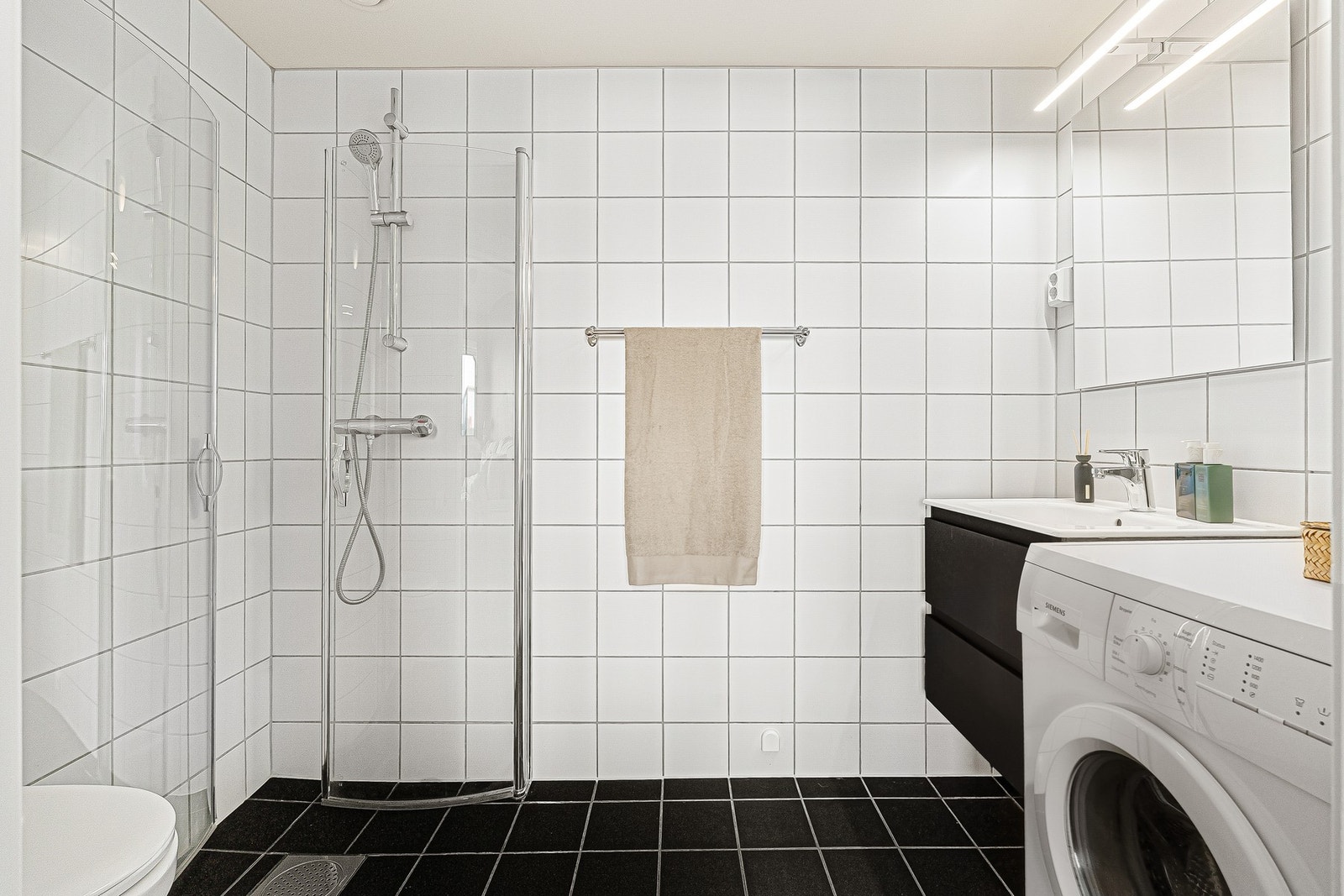 Flislagt og funksjonelt bad utstyrt med dusjhjørne, vegghengt toalett og praktisk opplegg for vaskemaskin. Galleribilde