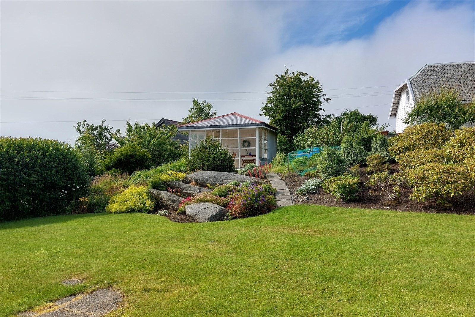Hagen er frodig opparbeidet med trær, plen busker og bed. Hellelagt gangsti til sommerstuen Galleribilde
