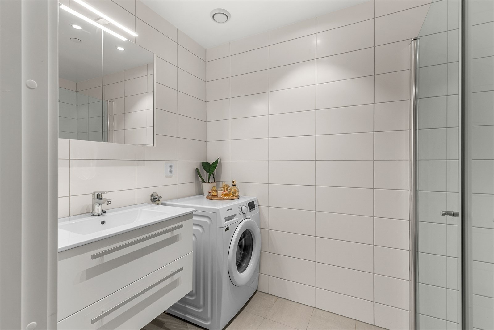 Lekkert og flislagt bad med rikelig med skap- og benkeplass samt et stort speil. Badet har behagelig gulvvvarme. Det er utstyrt med veggmontert wc, innfellbare dusjdører, servantskap, mekanisk avtrekk og opplegg for vaskemaskin. Galleribilde