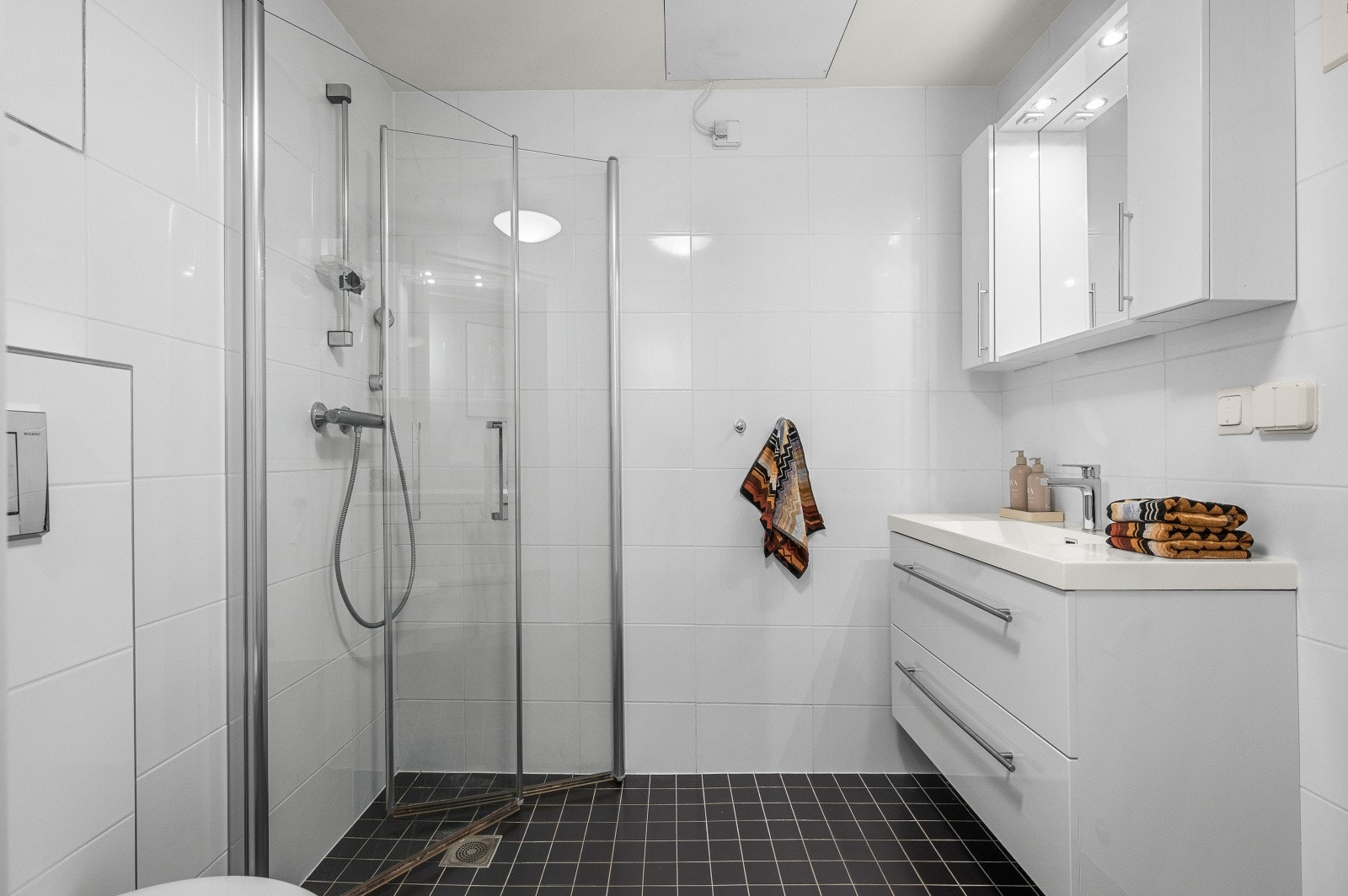 Moderne baderom utstyrt med veggmontert toalett, dusjhjørne, servant i servantskap og opplegg for vaskemaskin. Galleribilde