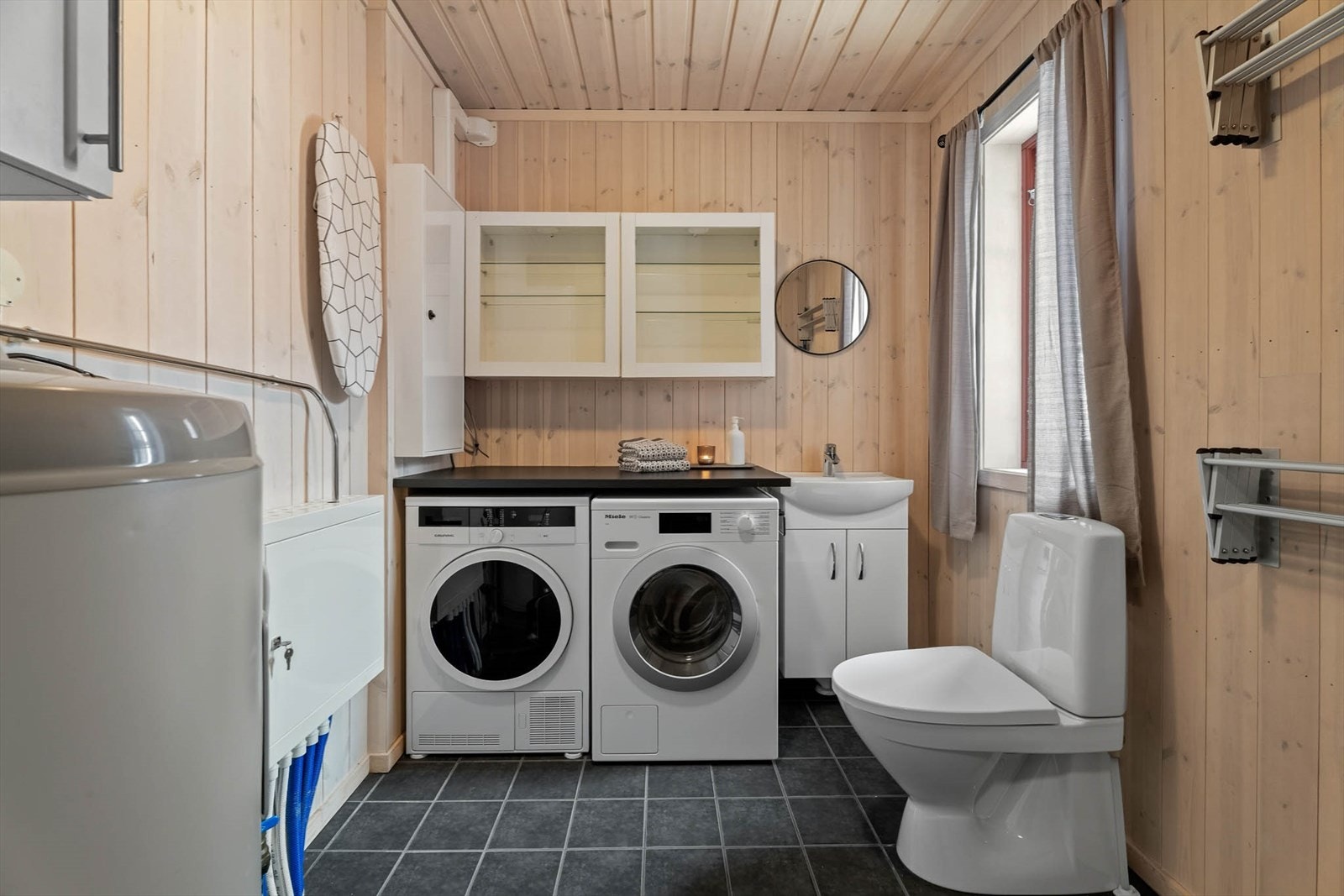 Vaskerommet har flis på gulv og panel på vegger. Her er det opplegg for vaskemaskin, benkeplate og overskap, samt 200l varmtvannstank.
I tillegg toalett og innredning med heldekkende hvit vask, underskap med slette fronter og speil. Naturlig ventilasjon. Galleribilde