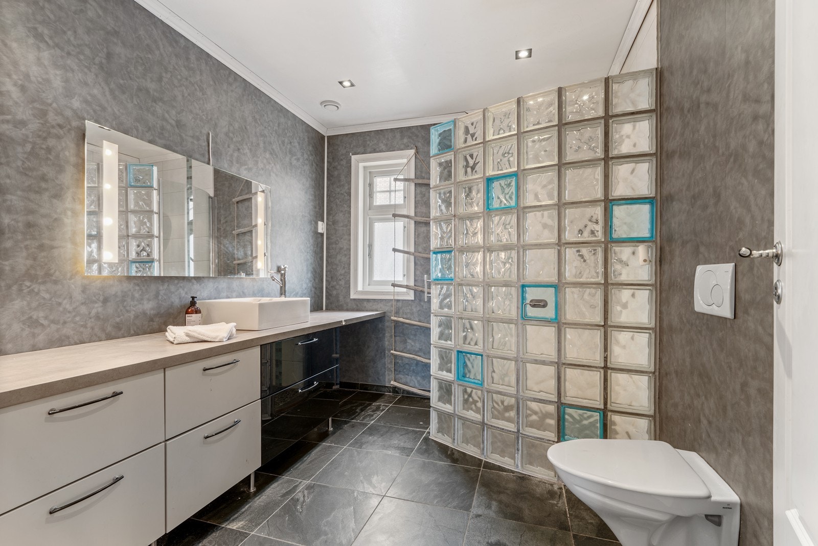 Baderom utstyrt med innredning med påliggende servant, veggmontert toalett, dusjhjørne med glassvegger og opplegg for vaskemaskin Galleribilde