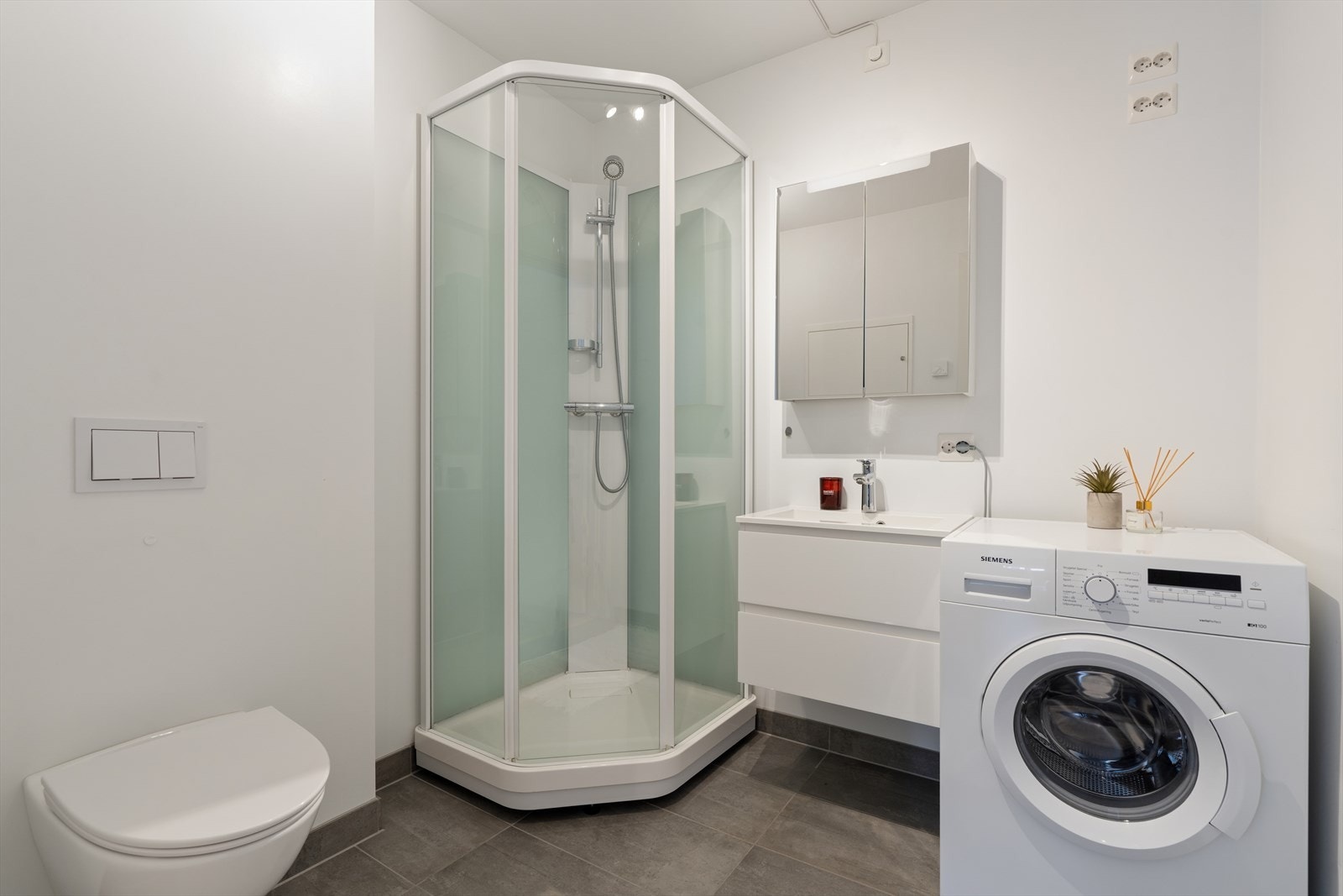 Badet har flislagt gulv og malte vegger. Rommet er innredet med vegghengt toalett, dusjkabinett, baderomsinnredning med servant og speilskap, samt opplegg for vaskemaskin. Galleribilde