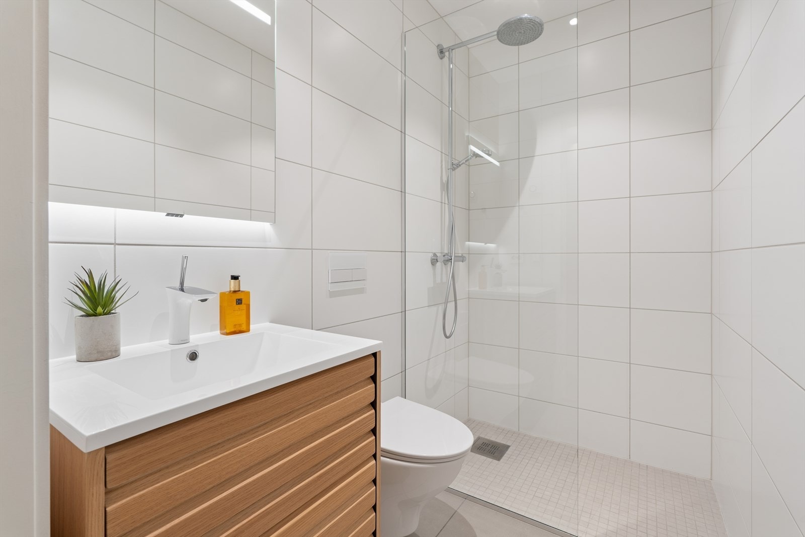 Baderommet i 1. etasje er flislagt, utstyrt med vegghengt toalett, dusjhjørne med glassvegg, baderomsinnredning med servant og speil. Galleribilde