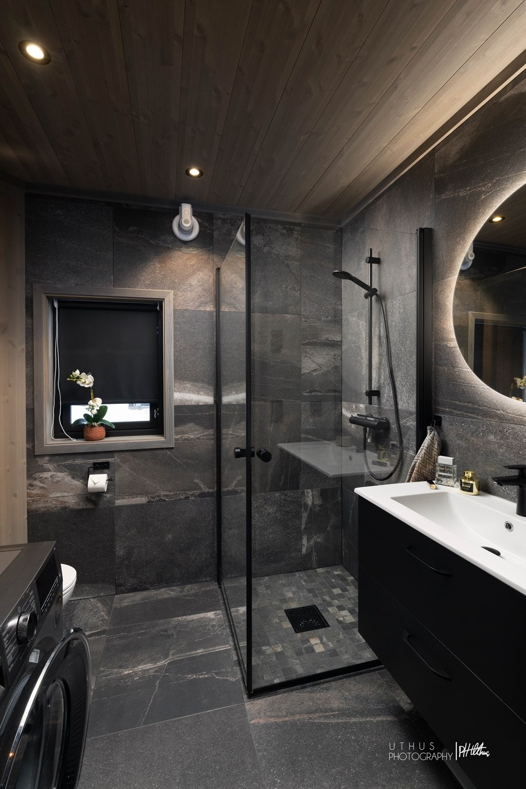 Delikat, flislagt bad med mørke overflater, dusjhjørne og opplegg for vaskemaskin. Galleribilde