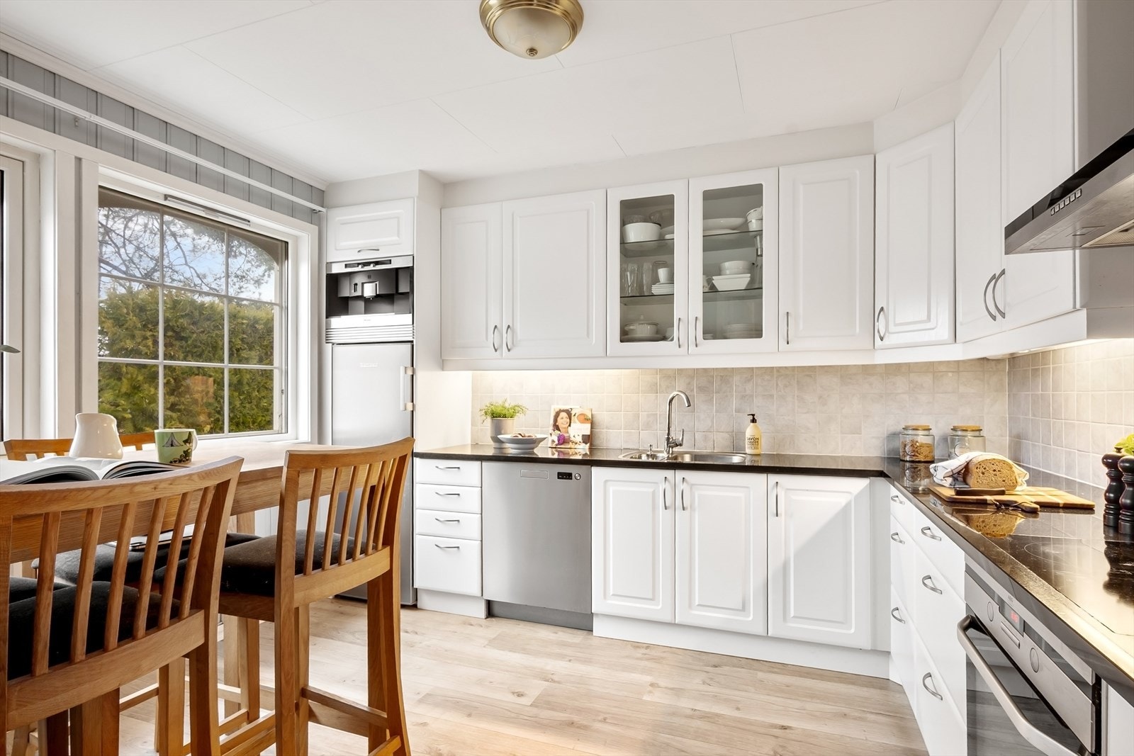 Rommet har store vindusflater og innredning fra 2015 med profilerte fronter, mørk benkeplate og integrert kaffemaskin over kjøleskap. Galleribilde