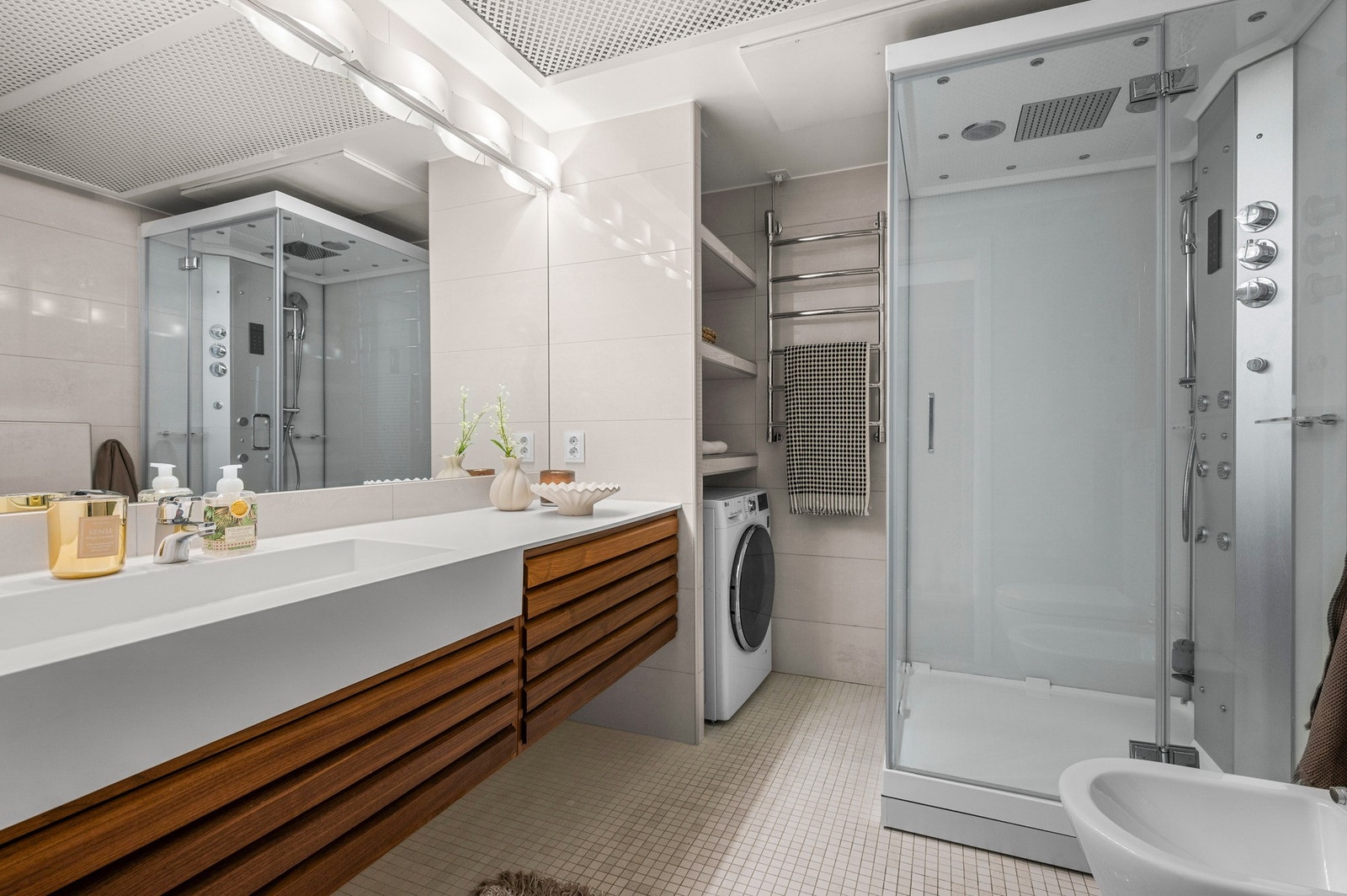 Badet er utstyrt med vegghengt toalett og bidet, servant og servantskuffer, dusjkabinett, oppheng for håndklær med varme, et stort speil med lys over, samt opplegg for vaskemaskin med hyller for praktisk oppbevaring. Galleribilde