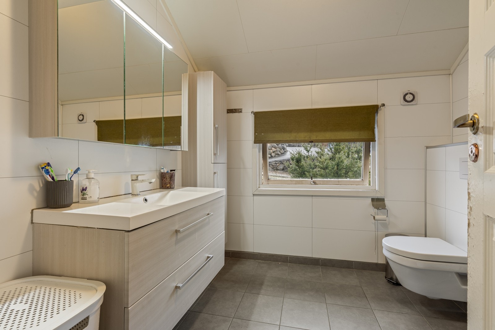 Badet er utført med fliser på gulv-og veggflater. Det inneholder dusjkabinett, servant i møblement, speilskap, vegghengt skap og vegghengt toalett. Galleribilde