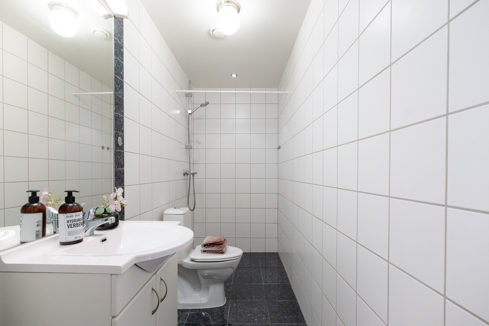 Badet i tilknytning til entréen har også fliser på gulv og vegger. Det er innredning med skap og ovenpåliggende servant, samt speil med belysning over. Videre er det dusj og toalett. Galleribilde